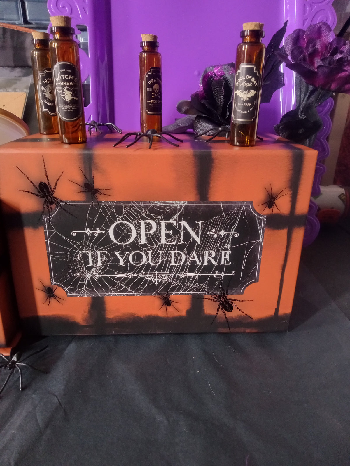 Halloween-themed box with spider decorations on a purple surface with bottles.